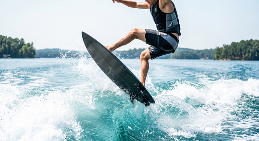 A wakesurfer mid-air above a boat wake on a sunny day, riding a black carbon fiber Smith Board Co. wakesurf board. His knees are tucked to his chest, demonstrating maximum vertical lift over the water.